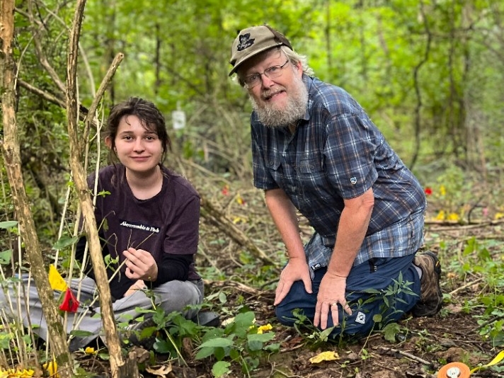Biology faculty and students sit in a vibrant, green woodland area, measuring honeysuckle growth.